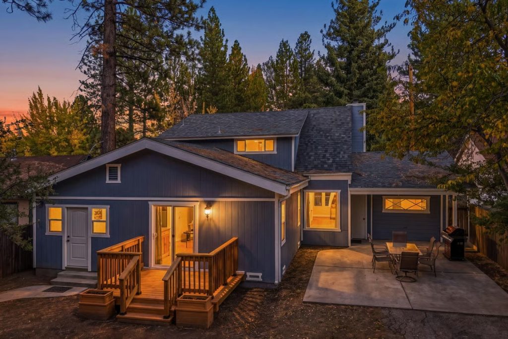 Exterior view of The Mountainaire Place cabin in Big Bear featuring a wooden deck, outdoor patio seating, and warm evening lights surrounded by tall pine trees.