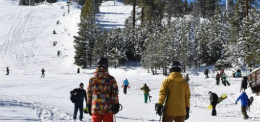 Two people holding snowboards in snowboarding gear looking up at a ski slope filled with riders in winter in Big Bear, California.