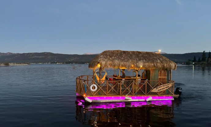 A tiki boat with colorful lights cruising on a calm lake during evening hours.