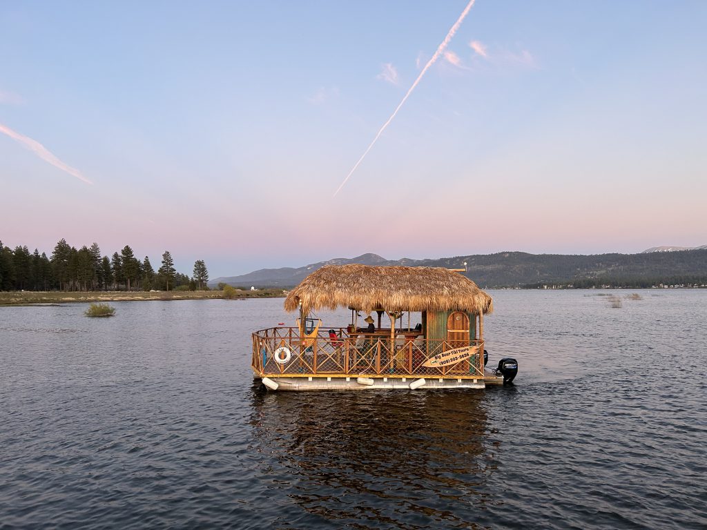 A tiki-themed pontoon boat with a thatched roof cruises on the calm waters of Big Bear Lake.