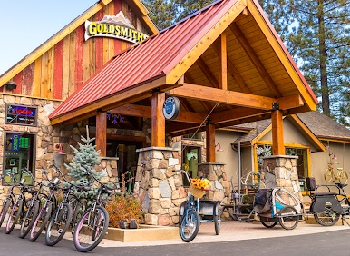 The exterior of a business called "Goldsmith's" in Big Bear, a rustic-style building with stone columns, a red metal roof, and several bicycles and bike trailers for rent or sale out front.