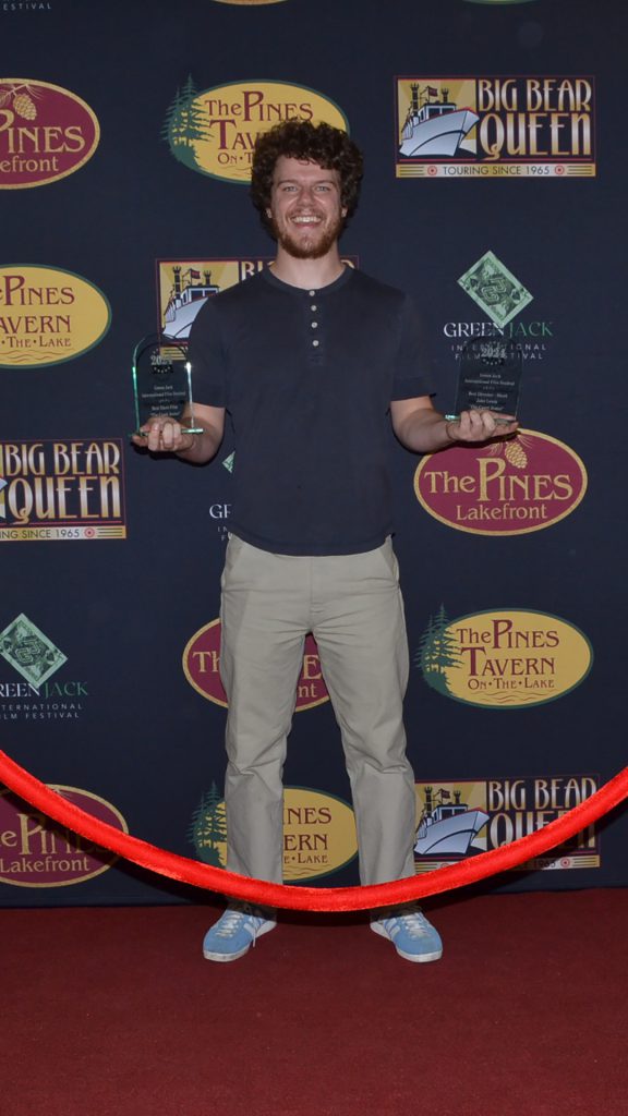 A young man holds two awards on a red carpet at a film festival in Big Bear.