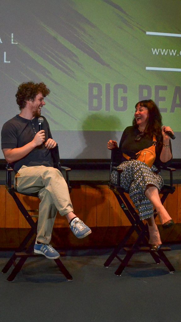 A man and a woman sit on director's chairs on a stage and laugh while holding a microphone at an event in Big Bear.
