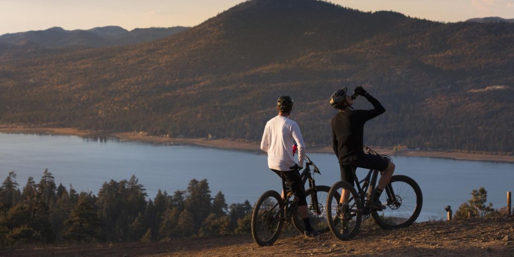 Two mountain bikers pause at a scenic overlook above Big Bear Lake, enjoying panoramic views of the lake and surrounding forested mountains during golden hour.