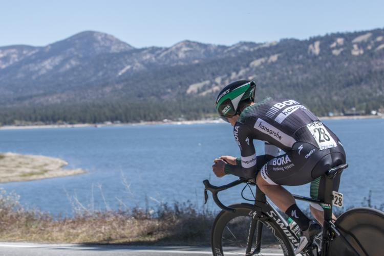 A road cyclist rides along the shoreline of Big Bear Lake with scenic mountain views in the background on a clear day.