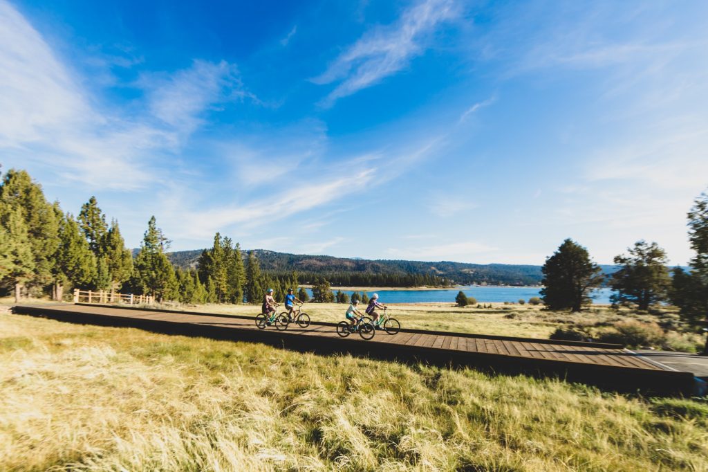 A group of cyclists riding on a scenic path near Big Bear Lake, surrounded by lush green fields and pine trees, with a bright blue sky above.