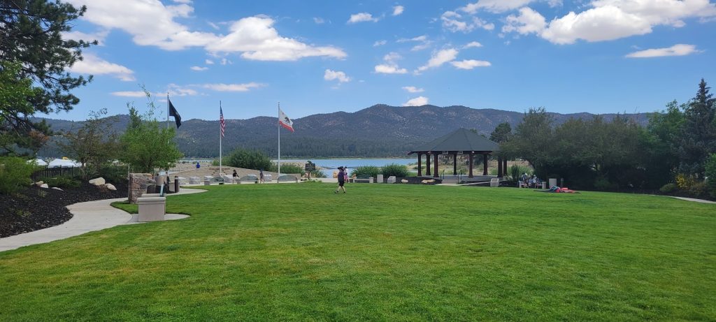 Wide view of Veterans Park in Big Bear featuring open green lawns, a memorial area with flags, a gazebo, and mountain views overlooking the lake.