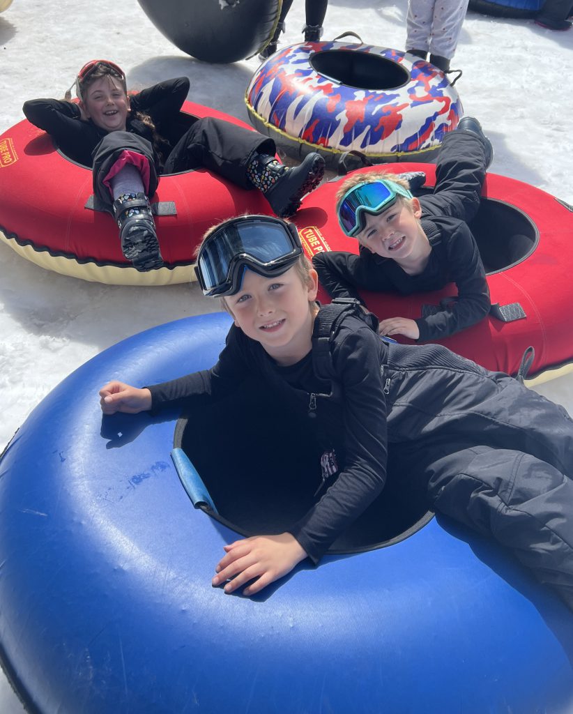 Three children in snow gear lie on large inflatable tubes on a snowy surface at Big Bear, with one child in the foreground smiling directly at the camera.