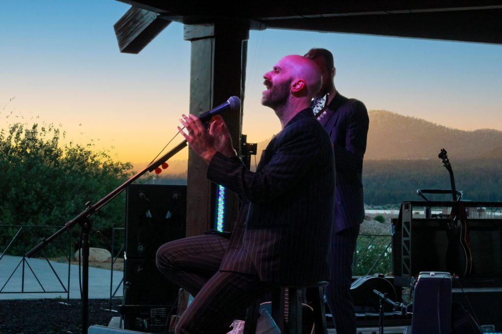 A musician with a shaved head and a beard sings with his hands clasped as another musician stands behind him, performing at an outdoor venue in Big Bear during sunset.