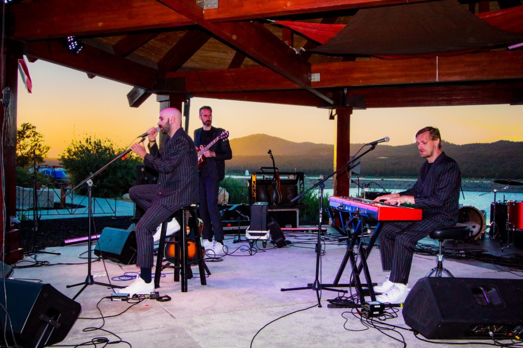 A three-man band, including a lead singer, a guitarist, and a keyboardist, performs on an outdoor stage with a lake and mountains in the background at sunset in Big Bear.