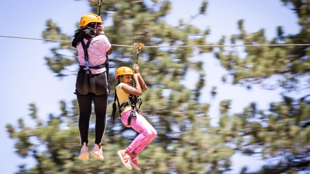 Two girls ziplining through a pine forest in Big Bear, California, enjoying a thrilling adventure high above the ground.
