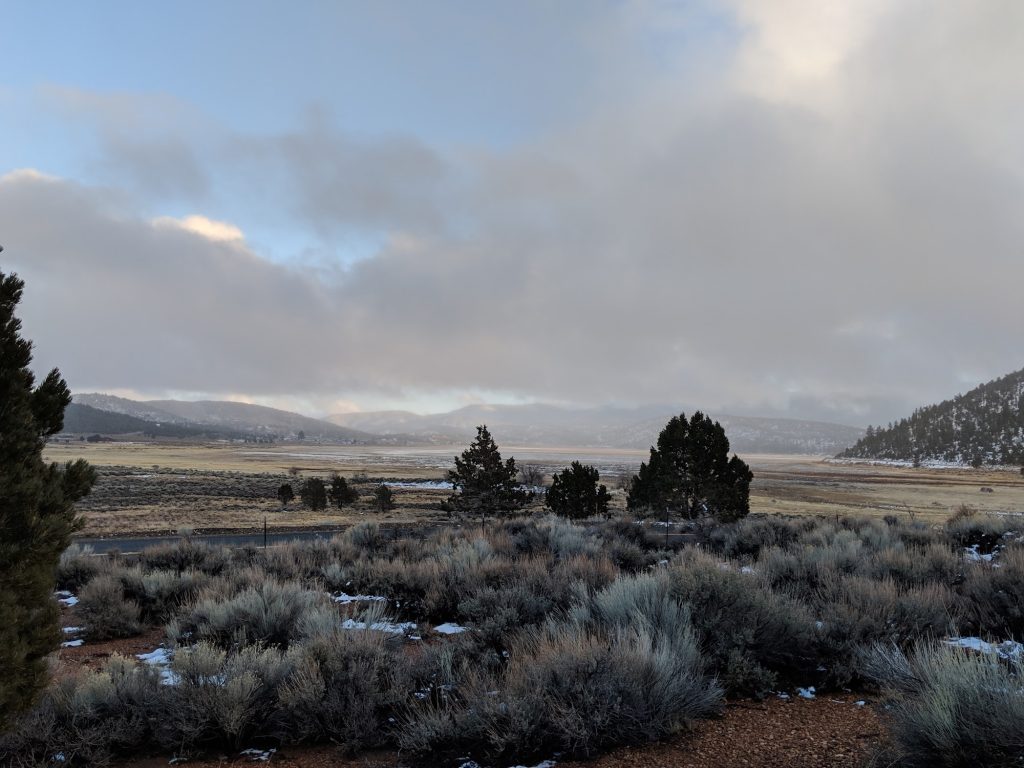 Scenic view of Baldwin Lake Ecological Reserve featuring sagebrush, patches of snow, open meadow, and surrounding mountains beneath dramatic winter clouds.