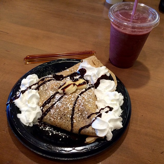 Plate with a folded crepe topped with banana slices, chocolate drizzle, whipped cream, and a berry smoothie on the side.