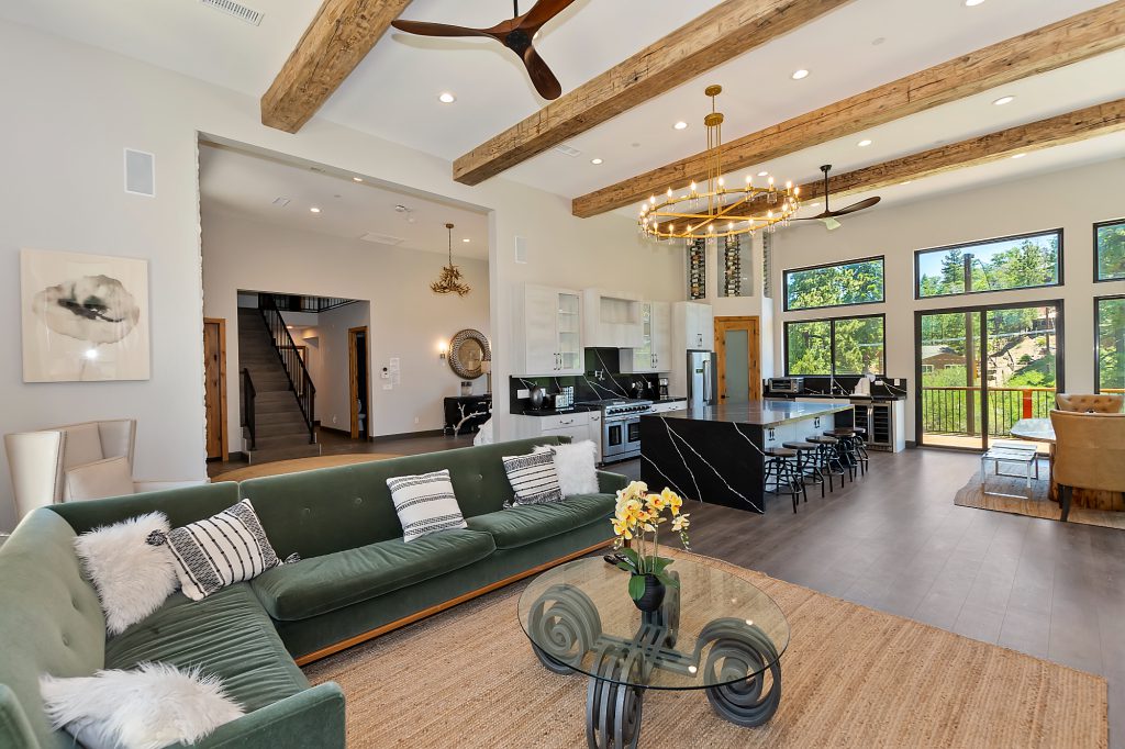 An open-concept cabin living area in Big Bear with a green velvet sectional sofa, a modern kitchen with a black marble island, and exposed wooden beams on the ceiling.