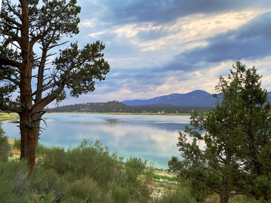 Scenic view of Big Bear Lake framed by pine trees with calm reflective waters, mountain backdrops, and dramatic clouds at sunset.