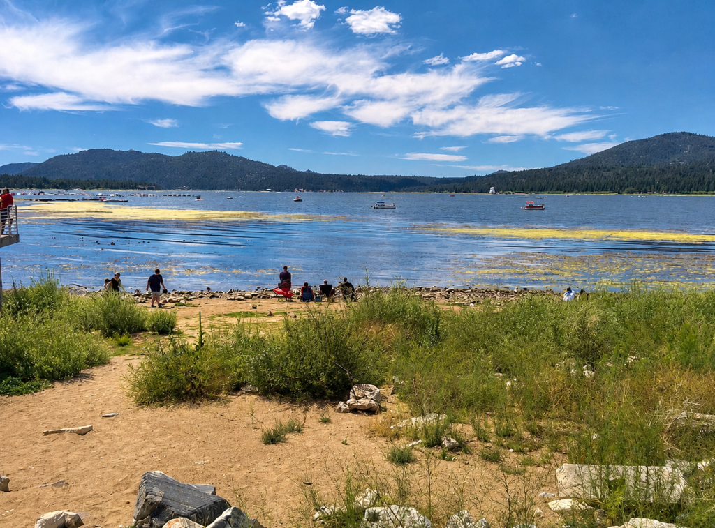 Visitors relaxing on rocky shoreline at Big Bear Lake with boats on water, grassy foreground, and scenic mountain landscape under blue sky