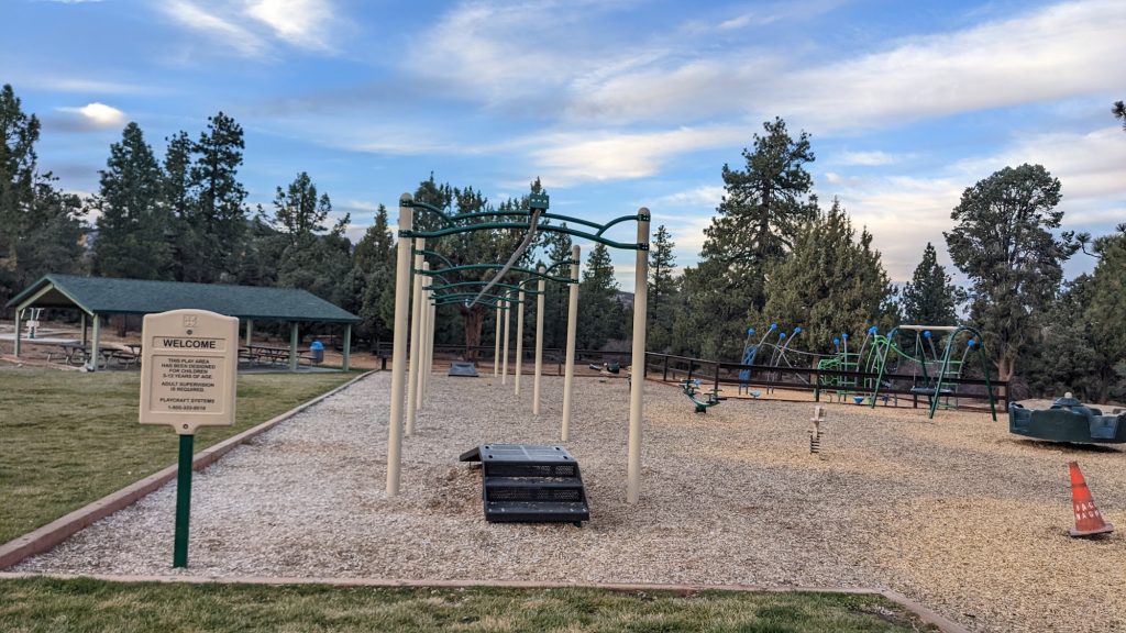 Outdoor park fitness and playground area featuring climbing bars, exercise structures, and picnic shelter surrounded by pine trees under a partly cloudy sky.