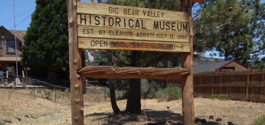 Wooden entrance sign for the Big Bear Valley Historical Museum displaying museum information and operating days, surrounded by pine trees and landscaped grounds.