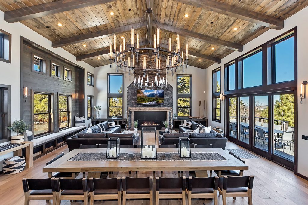 A grand and luxurious living room in a Big Bear cabin, with a large dining table, a stunning chandelier, a stone fireplace, and large windows with scenic views.