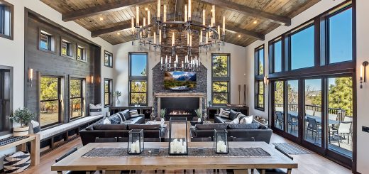 A grand and luxurious living room in a Big Bear cabin, with a large dining table, a stunning chandelier, a stone fireplace, and large windows with scenic views.
