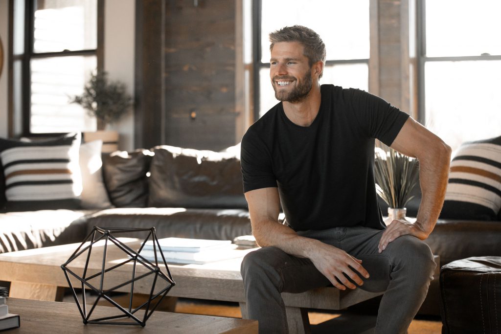 A smiling man in a black t-shirt and grey pants sits in a sunlit living room with a geometric sculpture on the coffee table.