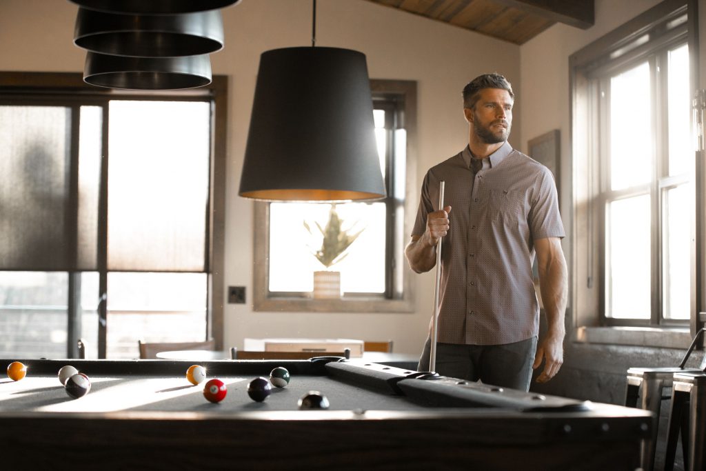 A man in a short-sleeved collared shirt stands by a pool table, holding a cue stick and looking out a large window.