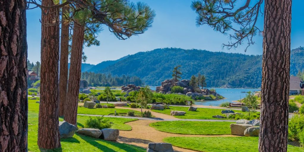 Scenic view of Boulder Bay Park in Big Bear featuring walking paths, pine trees, landscaped lawns, granite boulders, and the lake surrounded by mountains.