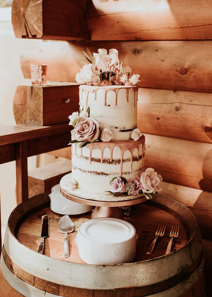 A two-tiered, rustic-style wedding cake with a pink drip glaze and floral decorations, displayed on a barrel at a restaurant in Big Bear.