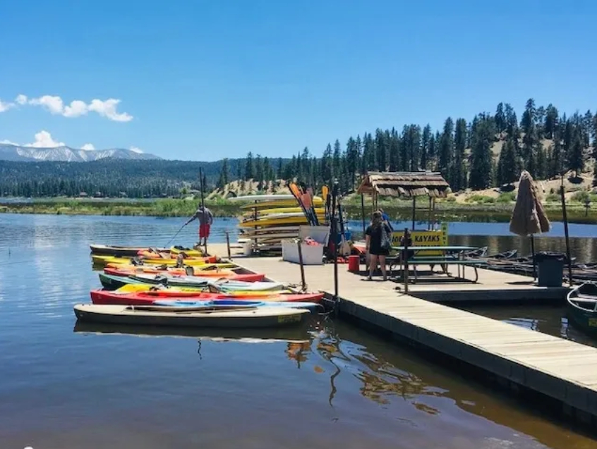Colorful kayaks and canoes lined up at a lakeside rental dock with staff station and scenic mountain lake background