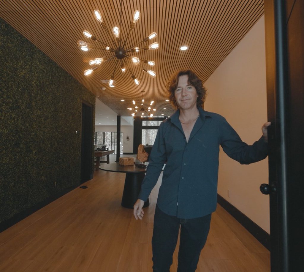 A man with wavy brown hair wearing a dark blue shirt and black pants is smiling while holding a door open, standing in the modern entryway of a Big Bear cabin with a slatted wood ceiling, a green moss accent wall on the left, and a unique starburst-style chandelier.