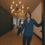 A man with wavy brown hair wearing a dark blue shirt and black pants is smiling while holding a door open, standing in the modern entryway of a Big Bear cabin with a slatted wood ceiling, a green moss accent wall on the left, and a unique starburst-style chandelier.