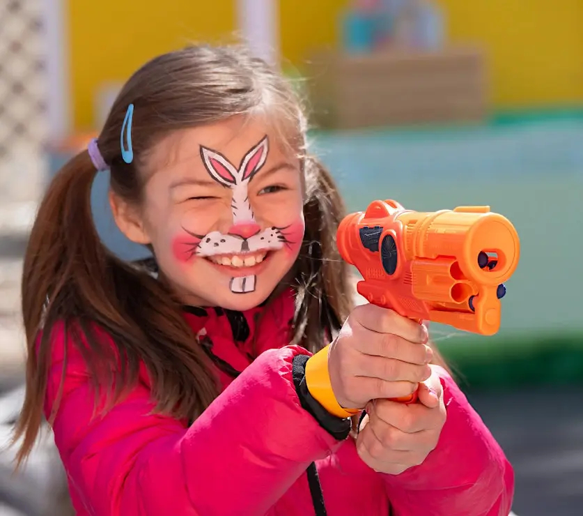 Child with bunny face paint holding toy blaster at Easter event