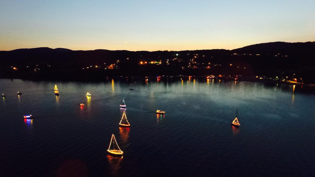 Illuminated boats are on the water at Big Bear during a boat parade with a town and mountains in the background.