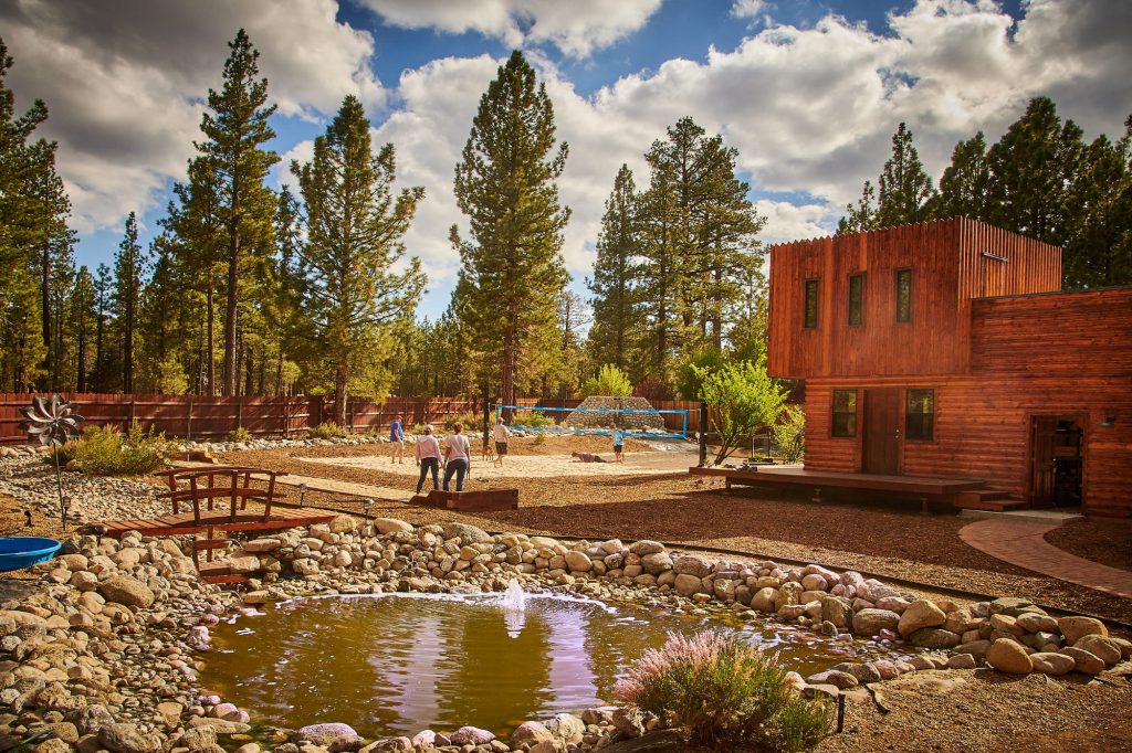 People are playing volleyball on a sand court next to a wooden building, with a small pond and fountain in the foreground, surrounded by tall pine trees in Big Bear.