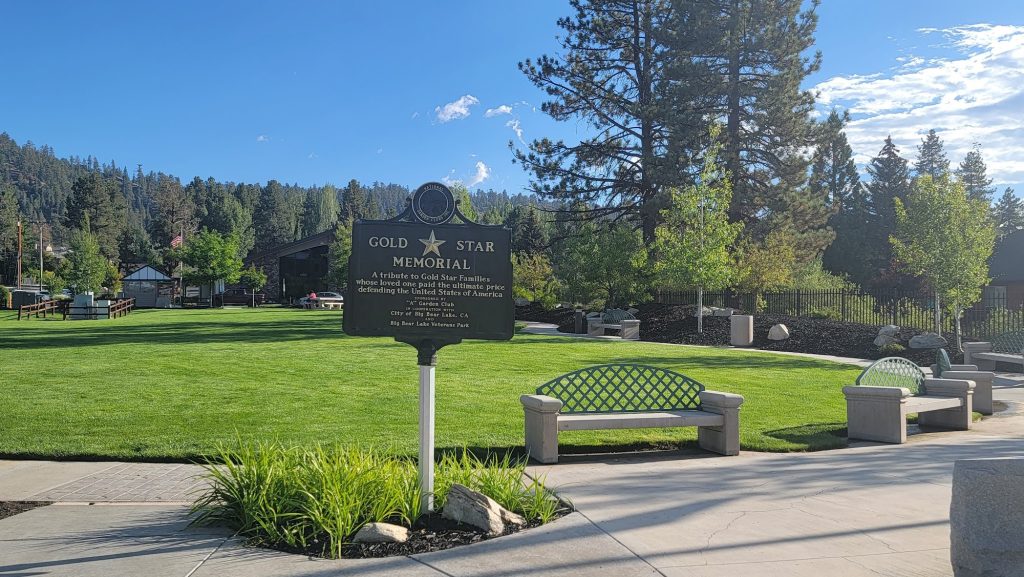 Gold Star Memorial sign at Big Bear Lake Veterans Park surrounded by green lawns, benches, and pine trees under a clear blue sky.
