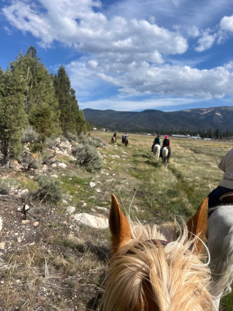 Horseback riders travel across a scenic meadow trail in Big Bear Lake with mountain views, pine trees, and a dramatic cloud-filled sky.