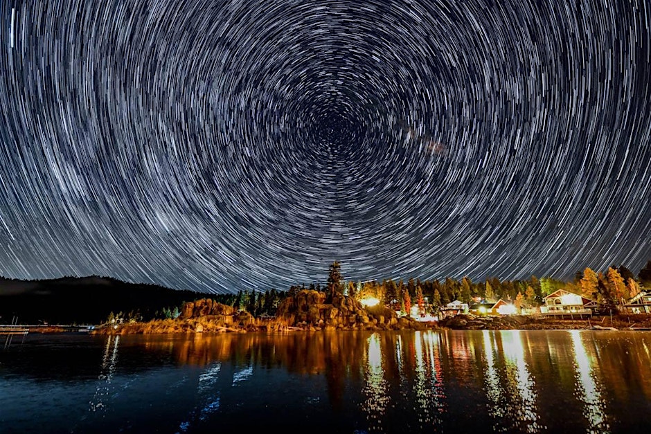 A night sky with star trails over a lake and trees in SkyFest Big Bear event.