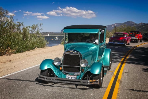 A line of vintage cars, led by a teal Ford Model A, drives along a road at Big Bear Lake.