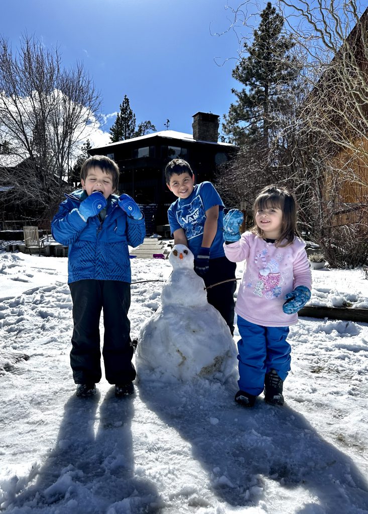 Three children pose with a snowman they built in a snowy yard at Big Bear.