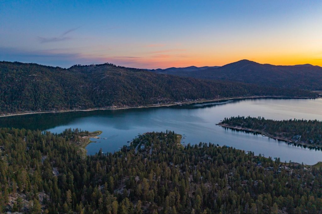 Aerial view of Big Bear Lake at sunset, showing forested hills and calm water reflecting the colorful sky.