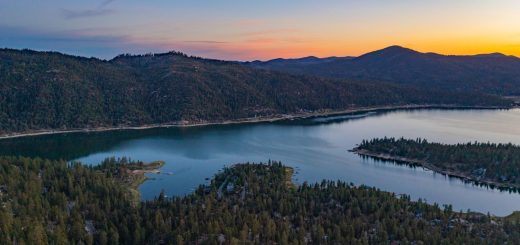 Aerial view of Big Bear Lake at sunset, showing forested hills and calm water reflecting the colorful sky.