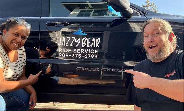 Two people are smiling and pointing at the Jazzy Bear Ride Service logo on the side of a black vehicle.