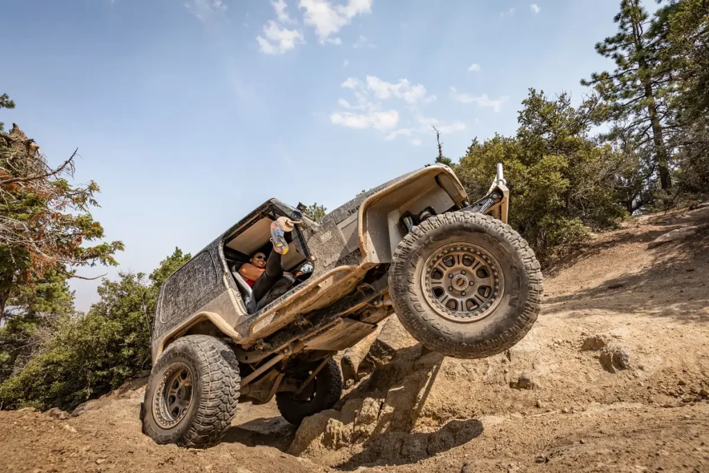 A dark jeep climbs a bumpy dirt road in Big Bear, with a driver visible inside, surrounded by trees and a blue sky.