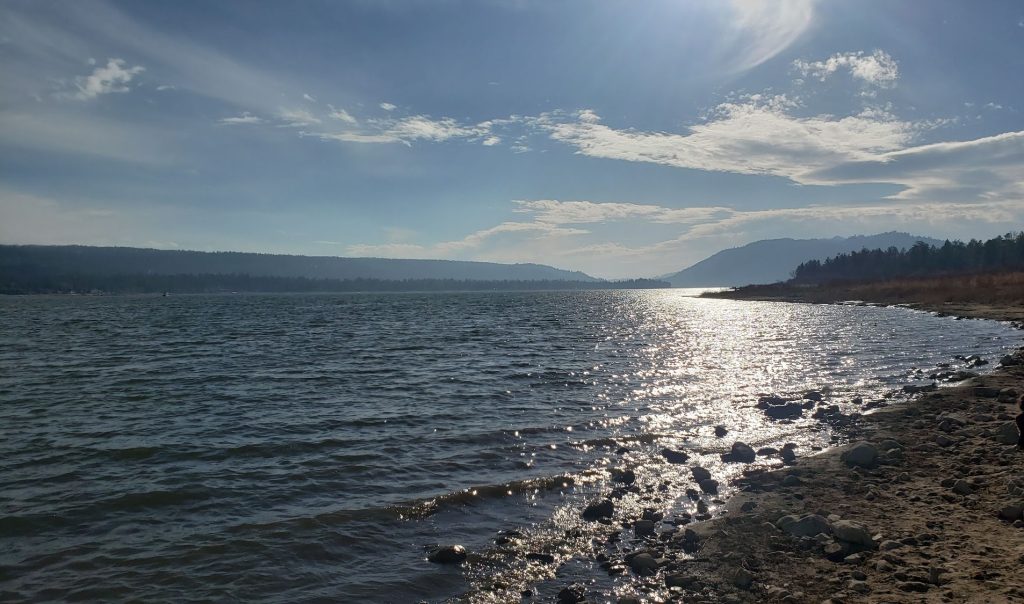 Scenic shoreline view from Juniper Point Picnic Area in Big Bear Lake featuring sparkling lake waters, a rocky beach, mountain silhouettes, and a bright afternoon sky.