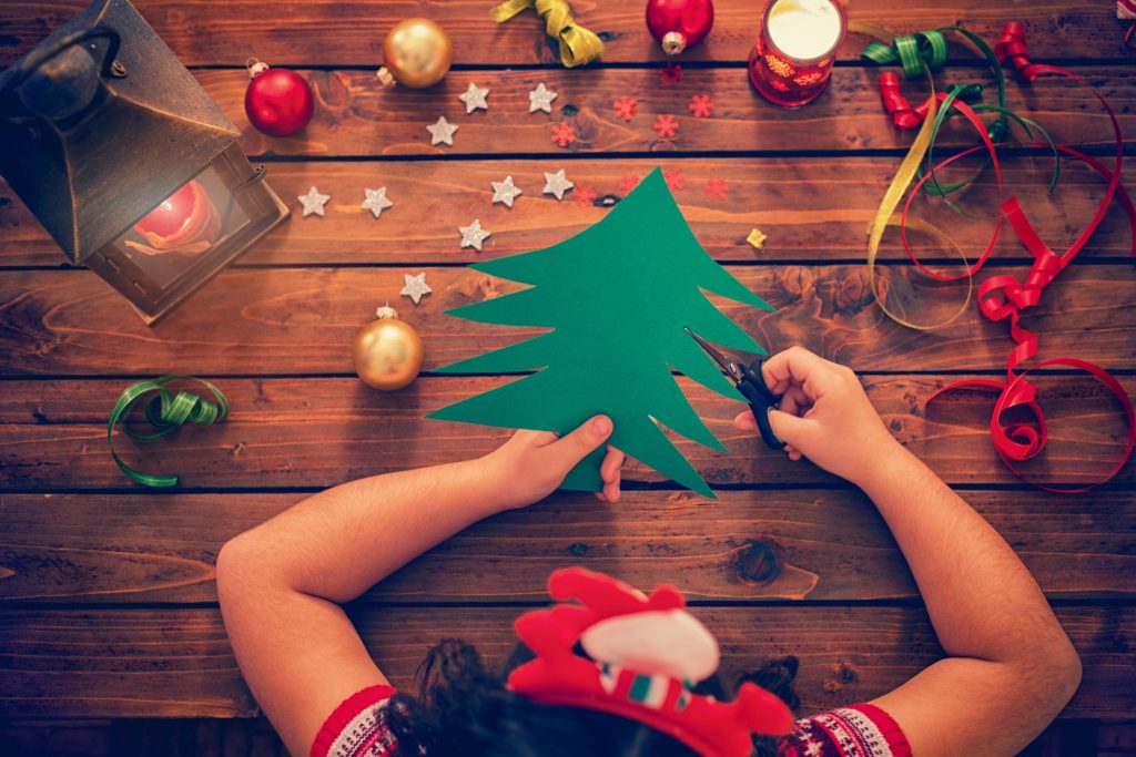 An overhead shot of a person wearing a Santa headband, cutting out a green paper Christmas tree on a rustic wooden table surrounded by Christmas ornaments, ribbons, and a lit lantern.