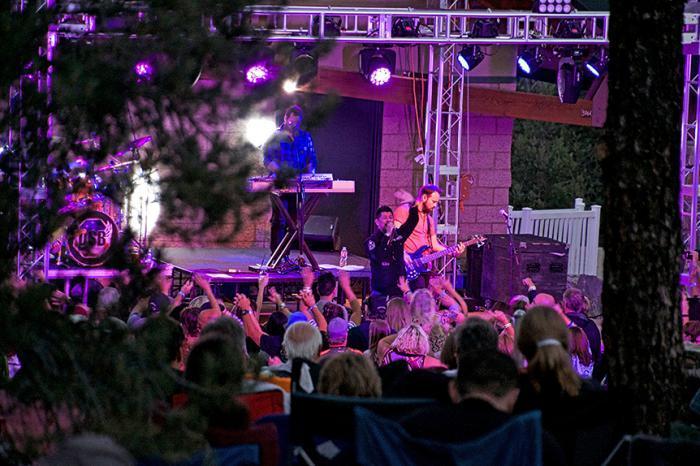 An outdoor concert scene with a crowd of people watching a band perform on a stage lit with purple lights at Big Bear