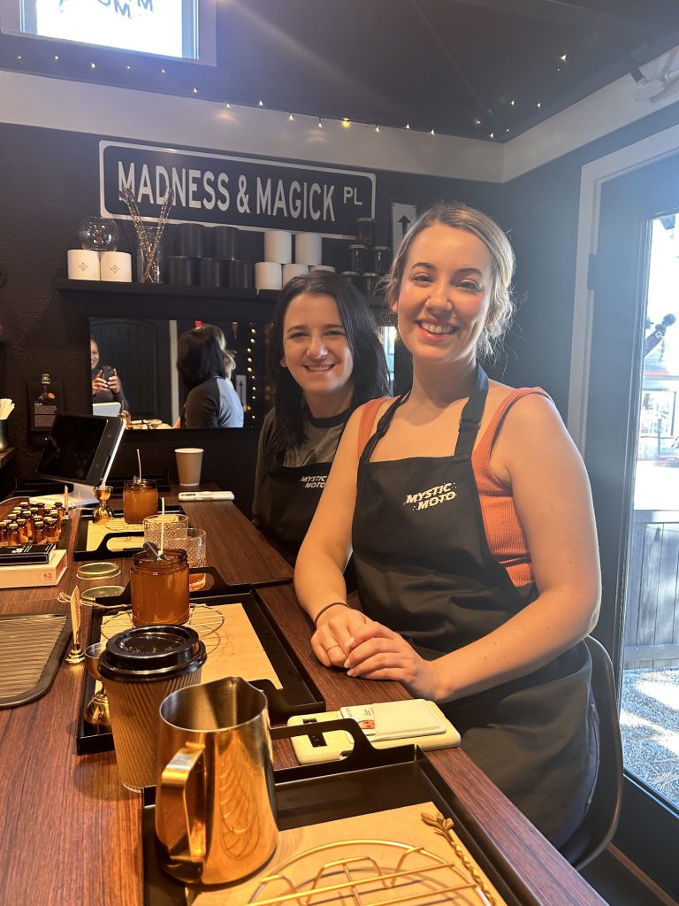 Two women smiling and posing at Mystic Moto, a candle-making store. The store features dark walls and various products in the background.