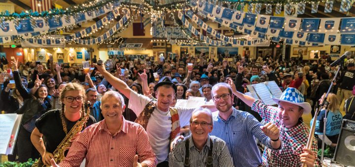 A band poses for a picture on a stage with a crowd of people in the background at the Big Bear Oktoberfest