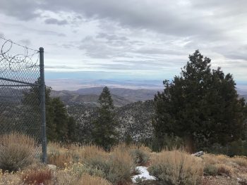Scenic mountain overlook with layered hills, trees, and distant desert landscape under cloudy sky.