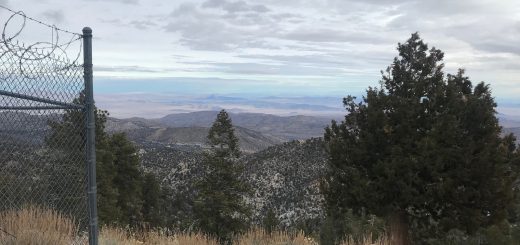 Scenic mountain overlook with layered hills, trees, and distant desert landscape under cloudy sky.
